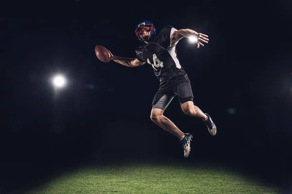 Jogador de futebol americano pulando com bola sob holofotes em preto — Fotografia de Stock