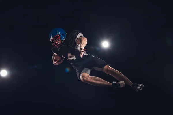 Vue du bas du joueur de football américain sautant avec le ballon sous les projecteurs sur noir — Photo de stock