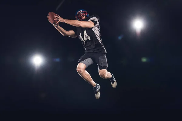 Вид снизу американского футболиста, прыгающего с мячом под прожекторами на черном — стоковое фото