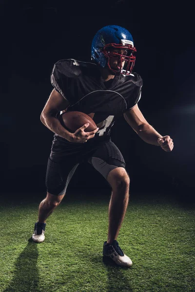 American football player holding ball and running on field on black — Stock Photo