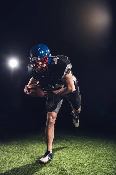 American football player holding ball and running on field under spotlights on black — Stock Photo