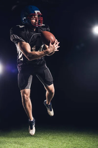 Jovem jogador de futebol americano saltando com bola sobre grama verde sob holofotes em preto — Fotografia de Stock