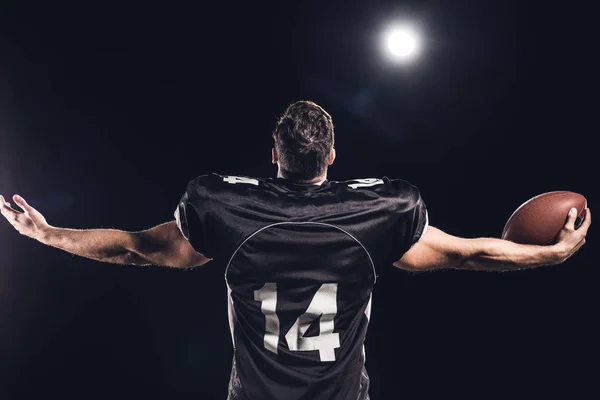 Visão traseira do jogador de futebol americano com bola olhando para cima com as mãos estendidas sob holofotes no preto — Fotografia de Stock