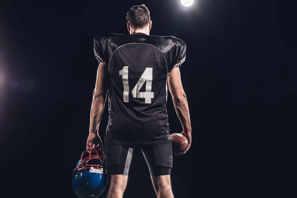 Visão traseira do jogador de futebol americano com bola e capacete sob holofotes em preto — Fotografia de Stock