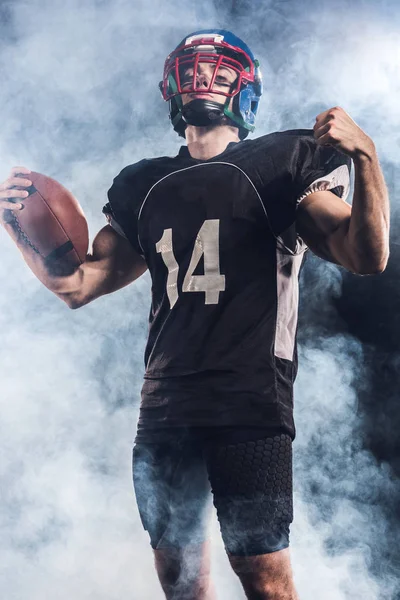 Bottom view of american football player with ball raisin fist against white smoke — Stock Photo