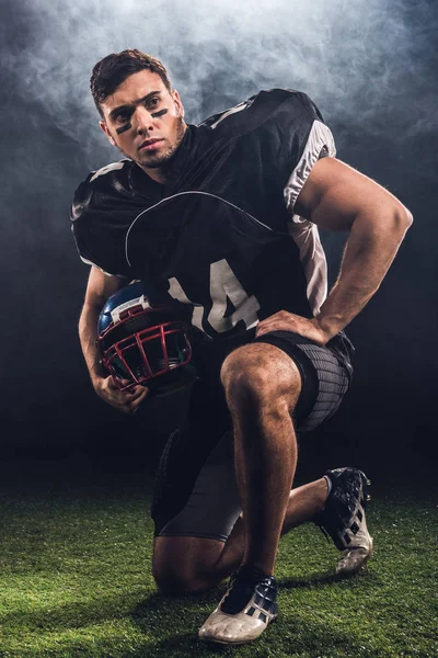 Attractive young american football player with helmet standing on knee on black — Stock Photo