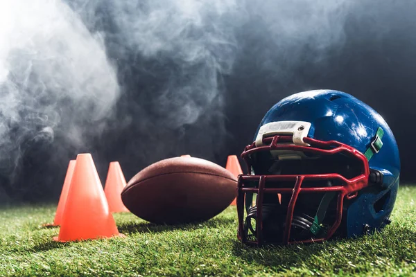Close-up shot of american football helmet with cones and ball on green grass with white smoke above — Stock Photo