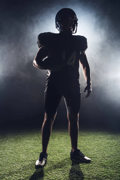 Silhouette di giocatore di football americano attrezzato con palla in piedi su erba verde contro il fumo bianco — Foto stock