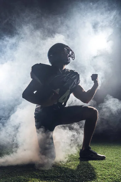 Young american football player standing on knee on green grass and raising fist in white smoke — Stock Photo