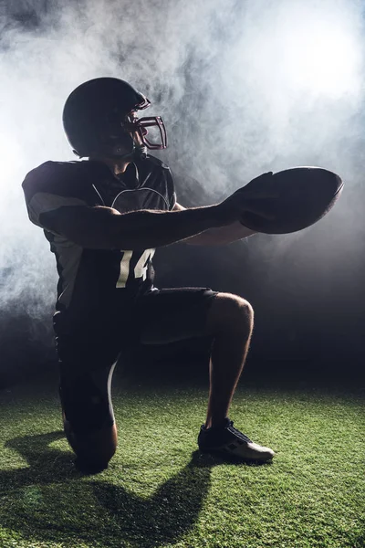Young american football player standing on knee on green grass and holding ball against white smoke — Stock Photo