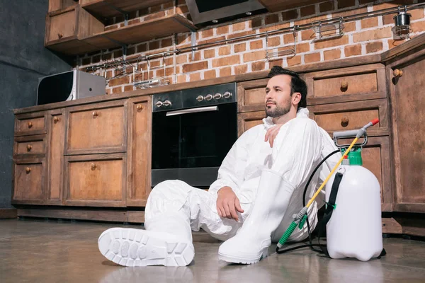 Trabajador cansado de control de plagas sentado en el suelo en la cocina - foto de stock