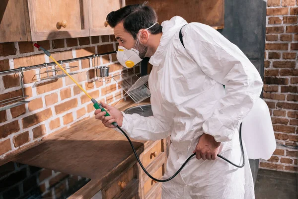 Vista lateral del trabajador de control de plagas rociando pesticidas debajo de los estantes en la cocina - foto de stock