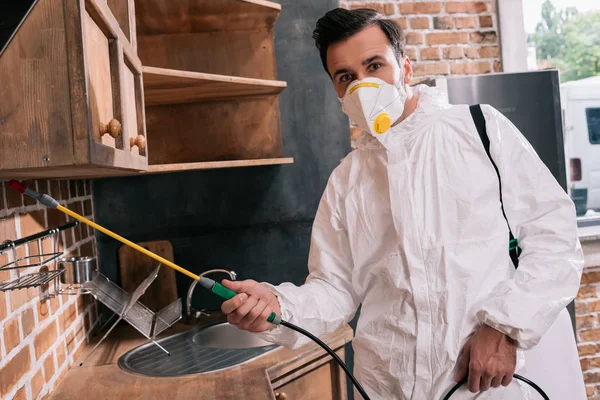 Trabajador de control de plagas pulverización de pesticidas debajo de los estantes en la cocina y mirando a la cámara - foto de stock