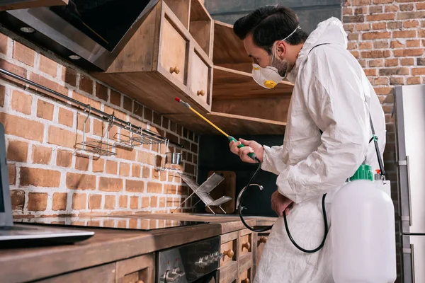 Vista lateral del trabajador de control de plagas rociando pesticidas debajo de los estantes en la cocina - foto de stock