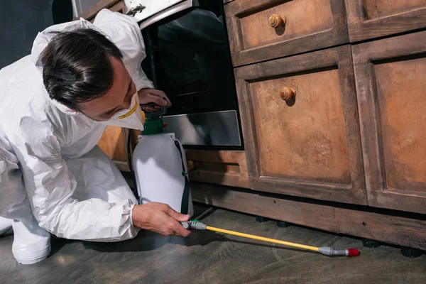 Pest control worker in uniform spraying pesticides under cabinet in kitchen — Stock Photo