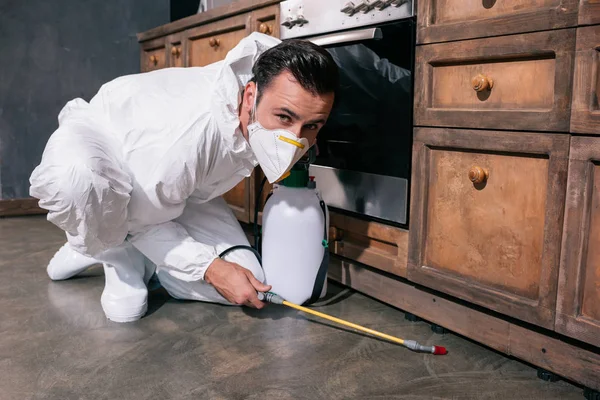 Trabajador de control de plagas pulverización de pesticidas en el suelo en la cocina y mirando a la cámara - foto de stock