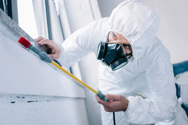 Pest control worker in respirator spraying pesticides under windowsill at home — Stock Photo