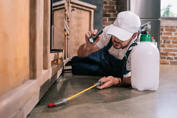 Pest control worker lying on floor and spraying pesticides under cabinet in kitchen — Stock Photo