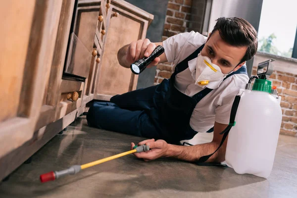 Pest control worker spraying pesticides under cabinet in kitchen and using flashlight — Stock Photo