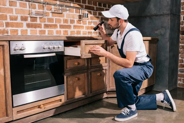 Trabajador de control de plagas examinando el gabinete en la cocina con linterna - foto de stock