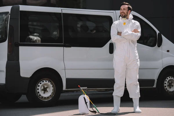 Travailleur souriant de la lutte antiparasitaire en uniforme debout avec les bras croisés près de la voiture et pulvérisateur sur la rue — Photo de stock