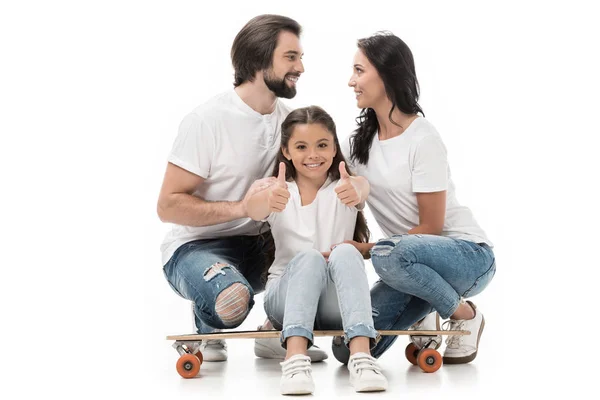 Smiling kid sitting on skateboard and showing thumbs up with parents behind isolated on white — Stock Photo