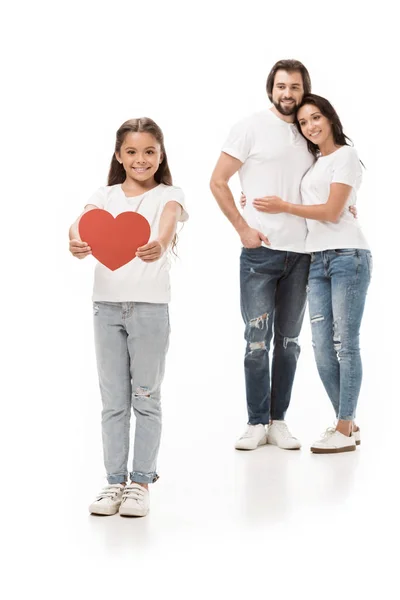Усміхнена дитина показує червоне паперове серце з батьками, обіймаючись позаду ізольовано на білому — стокове фото