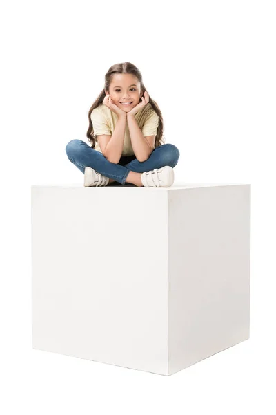 Lächelndes Kind in lässiger Kleidung sitzt auf weißem Würfel isoliert auf weißem — Stockfoto