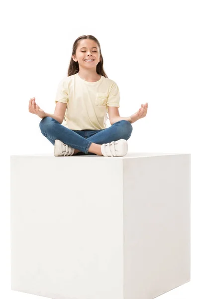 Enfant souriant en vêtements décontractés assis en pose de lotus sur cube blanc isolé sur blanc — Photo de stock