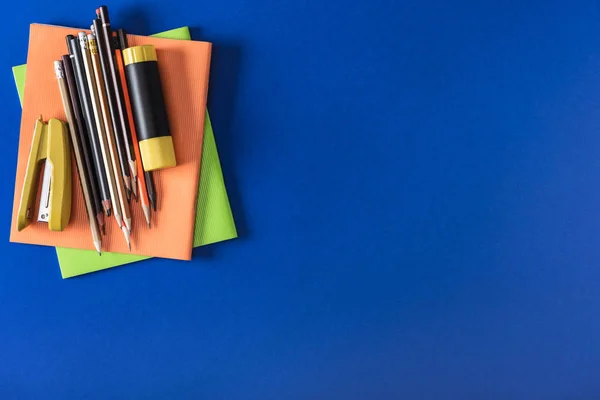 Vue du haut des manuels scolaires, différents crayons, agrafeuse et bâton de colle sur fond bleu — Photo de stock