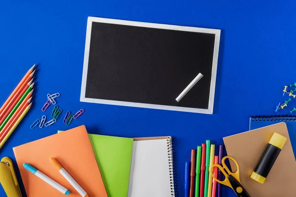 Top view of blank blackboard with chalk and variety stationery on blue background — Stock Photo