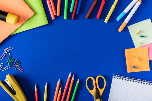 Draufsicht auf bunte Marker und Bleistifte, leeres Lehrbuch und diverse Papeterie auf blauem Hintergrund — Stockfoto