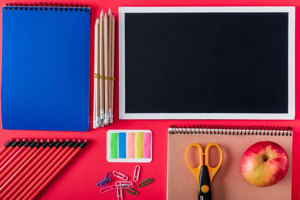 Top view of blank blackboard, apple and arranged variety stationery on red background — Stock Photo