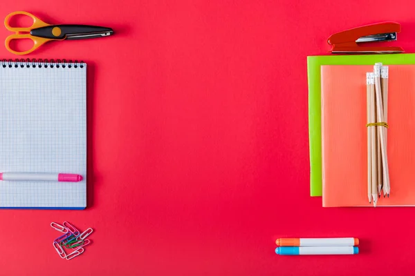 Top view of arranged textbooks, stapler, markers, paper clips, scissors and blank notebook on red background — Stock Photo