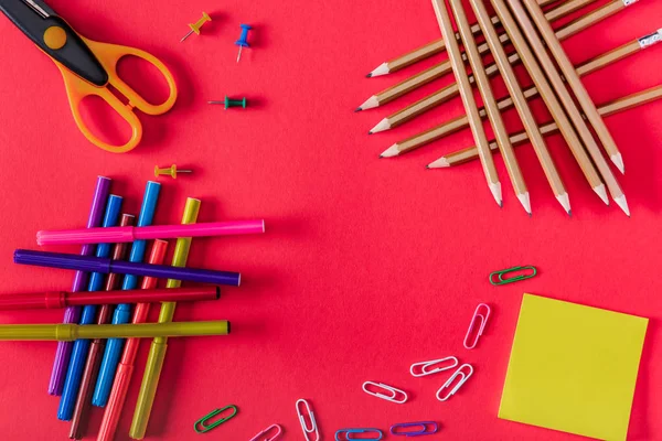 Вид зверху барвисті маркери, ножиці, шпильки, паперові затискачі, приклеїти його та олівці на червоному тлі — стокове фото