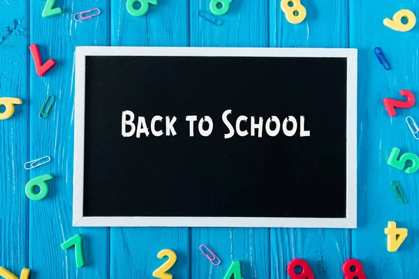 Top view of blackboard with lettering back to school, paper clips and colorful numbers on blue wooden background — Stock Photo