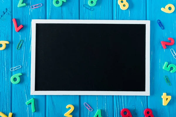 Draufsicht auf leere Tafel, bunte Zahlen und Büroklammern auf blauem Holzgrund — Stockfoto