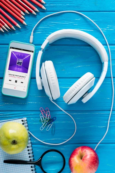 Ansicht des Smartphones mit Apple-Musikapplikation auf dem Bildschirm, Kopfhörer, Äpfel und Papeterie auf blauem Holzhintergrund — Stockfoto