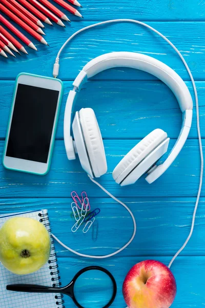 Вид сверху на наушники, смартфон с чистым экраном, яблоки и канцелярские принадлежности на синем деревянном фоне — стоковое фото