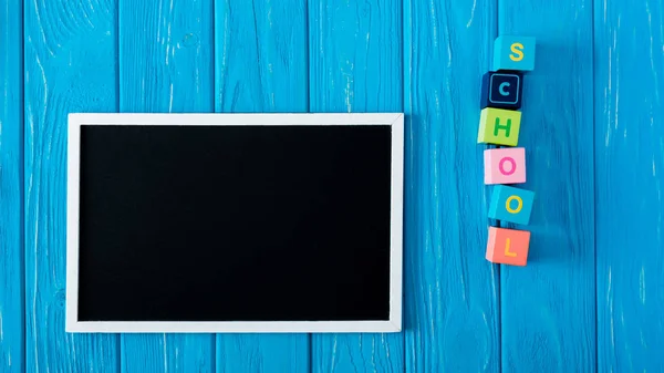 Top view of blank blackboard and lettering school made from cubes on blue wooden background — Stock Photo