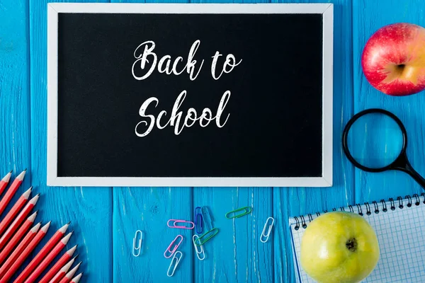 Top view of blackboard with lettering back to school, apples, blank notebook, pencils and paper clips on blue wooden background — Stock Photo