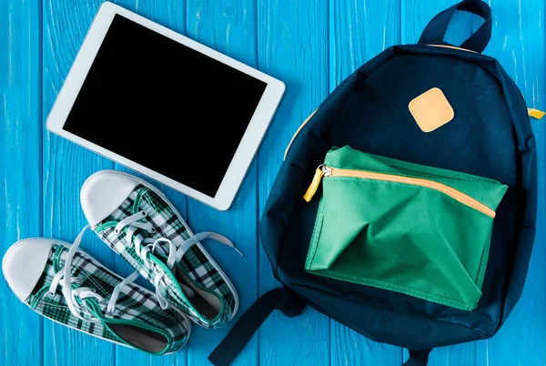 Vista superior de la tableta digital con pantalla en blanco, zapatillas de deporte y mochila sobre fondo de madera azul - foto de stock