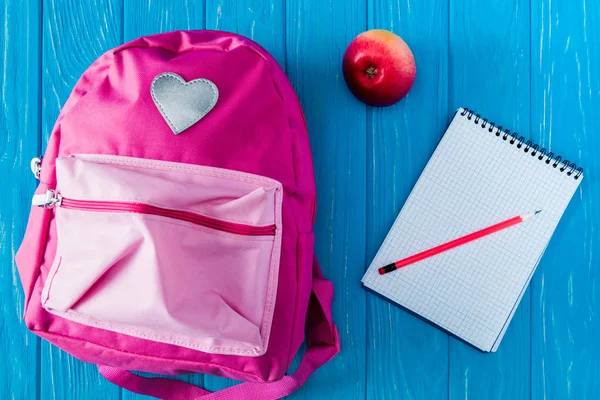 Vista superior de la manzana, mochila rosa, cuaderno en blanco y lápiz sobre fondo de madera azul - foto de stock