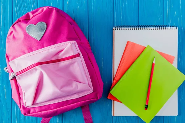 Vista superior de la mochila rosa, cuaderno en blanco, libro de texto y lápiz sobre fondo de madera azul - foto de stock