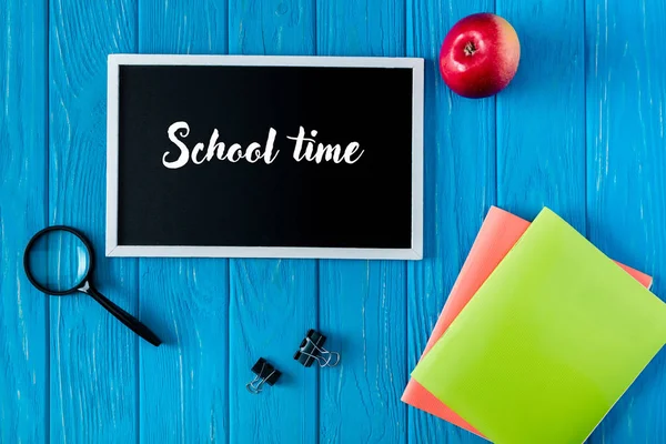 Top view of blackboard with lettering school time, apple and stationery on blue wooden background — Stock Photo