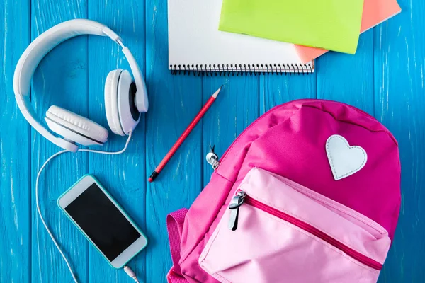 Vue du haut du sac à dos rose, smartphone avec écran vierge, écouteurs et manuels scolaires sur fond en bois bleu — Photo de stock