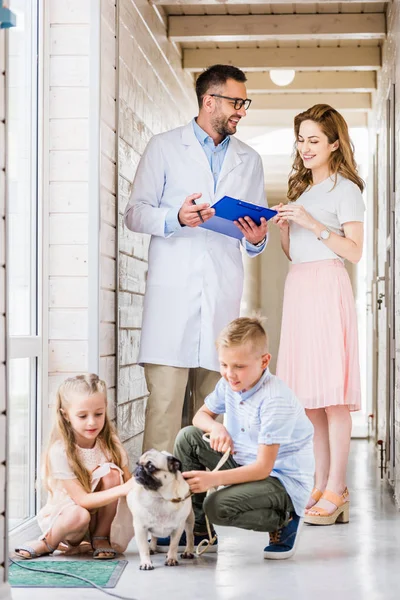 Tierarzt zeigt Frau etwas im Klemmbrett, Kinder spielen mit Mops in Tierklinik — Stockfoto