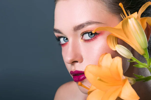 Крупным планом портрет стильной молодой женщины с модным макияжем и оранжевыми цветами лилии, смотрящей на изолированную на сером камеру — стоковое фото