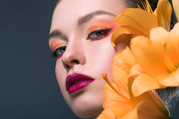 Nahaufnahme Porträt einer attraktiven jungen Frau mit modischem Make-up und orangefarbenen Liliumblüten, die isoliert auf grau in die Kamera blickt — Stockfoto