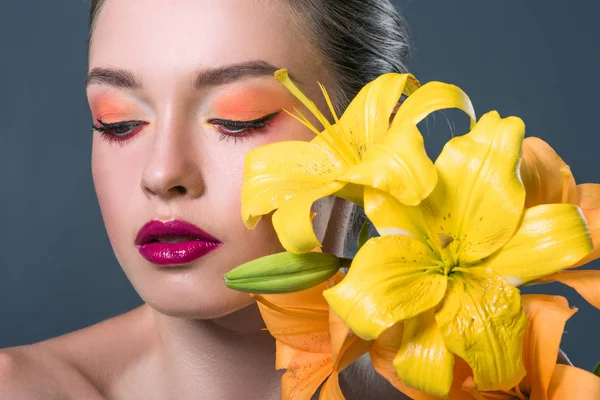 Ritratto ravvicinato di bella giovane donna con trucco alla moda e fiori di lilium gialli isolati su grigio — Foto stock